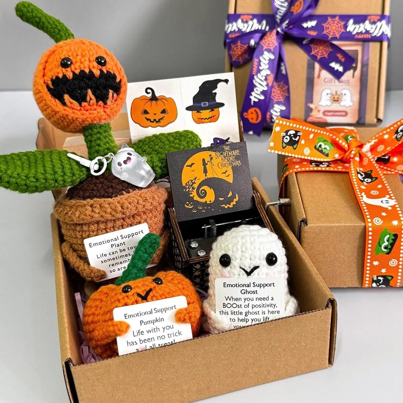 A Halloween-themed gift box with handmade crocheted items: a Jack-o'-lantern, ghost, and potted plant with smiley faces.